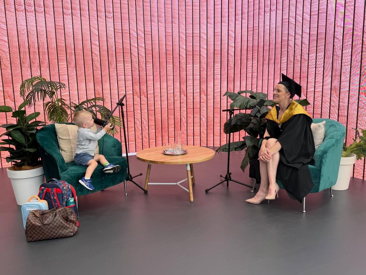 Tash Miller in her graduation gown on a podcast set, with Archie at the other microphone